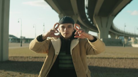 Zoom in shot of young hip hop dancer looking at camera and performing hand moves while dancing under urban bridge on sunny day