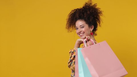 Charming cute young latin curly woman 20s wears casual flower dress holding package bags with purchases after shopping pointing on workspace area copy space mock up isolated on plain yellow background