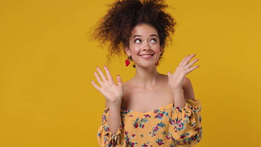 Vivid happy young latin curly woman 20s wears casual flower dress dance clenching fists waving rising expressive gesticulating hands fooling around have fun enjoy isolated on plain yellow background