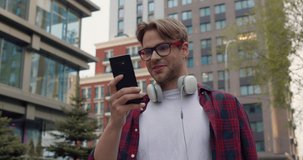 Smiling Guy Student in Eyeglasses recording Video Message using Smartphone. Happy Young Caucasian Man with Headphones on neck Walking at city street. Enjoys Online Conversation. Applications. Devices.