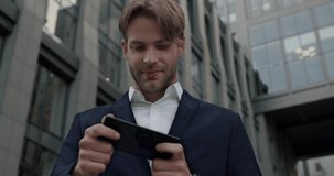 Exited Businessman playing Games on Smartphone near Office Center. Attractive Caucasian Entrepreneur using Mobile Applications. Spending Free Time from Work. Modern Device. Victory. Winner.
