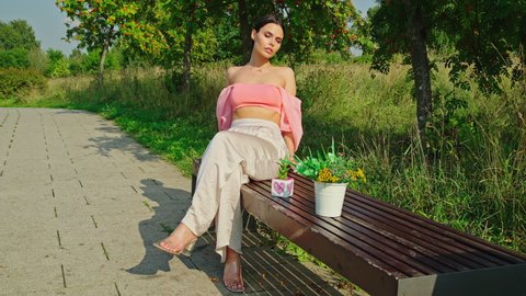 Attractive young woman sitting on bench and relaxing against trees in a park, forest or garden. Pots with flowers stay on bench in sunny day. Girl with naked shoulders stands under a rowan tree with c