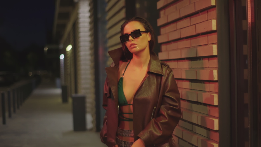 Party girl posing against modern night city. Fashion sexy woman holding match in her teeth. Beautiful hot model wearing brown jacket, green bra, sunglasses. 4K