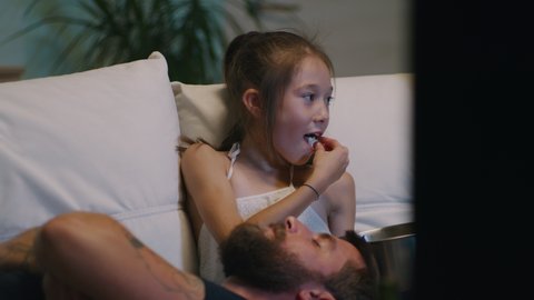 Girl eating and sharing popcorn with brother and Asian mother while sitting on couch near sleeping father and watching movie on TV in evening.