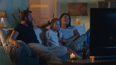 Mixed race girl entering living room  on sofa near multiracial mother and father watching TV at night at home