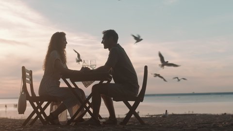 Long shot of silhouettes of happy man and woman sitting at table on sandy lakeshore, holding hands, talking, having wine at dusk, seagulls flying around
