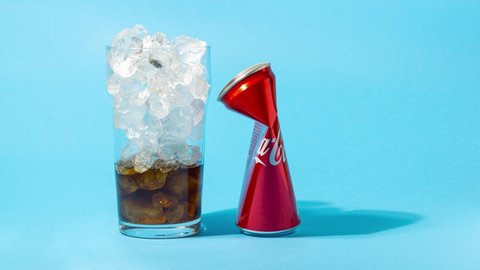UKRAINE, KIEV - SEPTEMBER 02, 2021: Glass of cola with ice is filled with Coca-Cola, the can is crumpling on blue background. Looped Stop motion animation