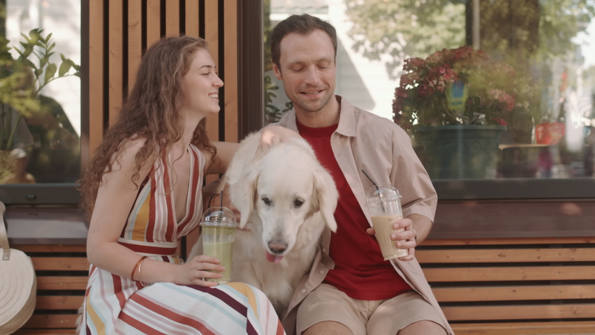Medium long of joyous Caucasian woman and man sitting outdoors coffee shop, holding iced coffee and matcha, petting their adorable dog Royalty-Free Stock Footage #1078573880