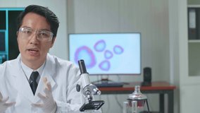 Asian Scientist In The Laboratory With A Microscope And Speaking To The Camera
