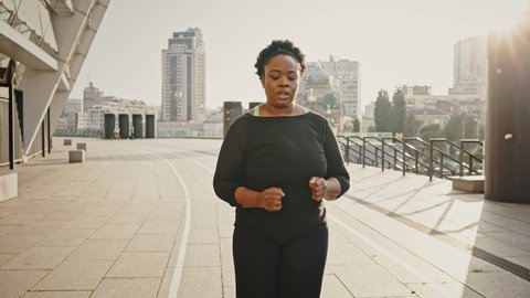 Hard slimming process. Outdoor portrait of young overweight african american woman running alone in morning, feeling tired of cardio training, slow motion