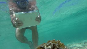 Slow motion of a man in snorkel underwater using pad in water-proof case to take pictures or shoot video of beautiful coral reef