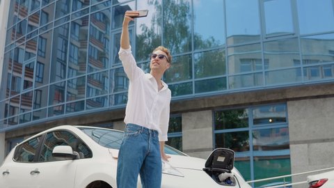 Stylish modern young curly man stands near an electric car charging and takes a selfie with his smartphone