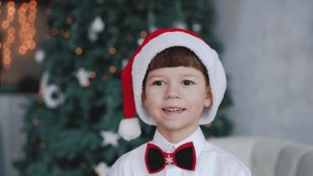 Close-up view 4k video portrait of cute happy smiling handsome little boy wearing red holiday Santa hat while sitting in Christmas interior