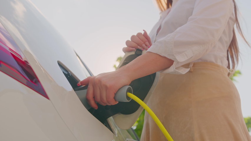 Low angle shot of a young smiling Caucasian woman opening an electric car charging socket cap and plugging in a charger | Shutterstock HD Video #1078593869