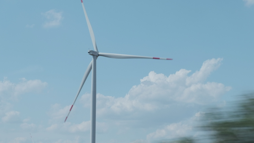 The view from the car window of the flickering tree branches against the rotating blades of a wind turbine generator at a wind farm. Royalty-Free Stock Footage #1078597316