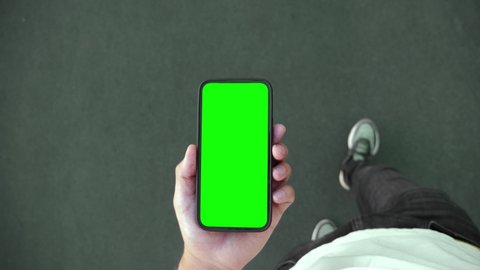 Young Person Walk and Use Green Screen Chroma Key Smartphone Top Point of View. Caucasian Man Hold in Hand Phone Mock-up and Watching Video Call, Copy Space Browsing. Go in Urban Cellphone Close-up 4k