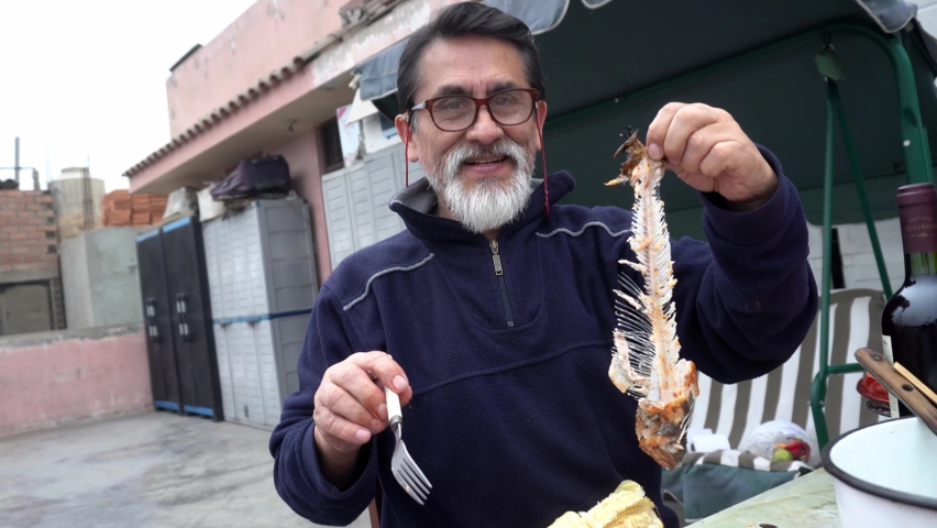 an elderly man with glasses holding a meatless trout fish with only bones proud of his cooking on a family rooftop grill in the daytime in 4K
