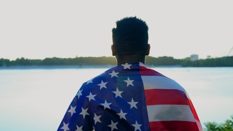 Back view afro-american man ethnic black with American flag on the shoulders looks into distance at sunrise in summer. African male stands on beach coast river with USA flag. 4th july independence day
