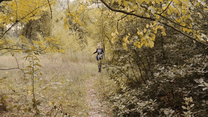 A girl with a scary make-up in a skeleton costume, a mask with rhinestones in a gloomy autumn forest. Runs along the path jumping merrily. Halloween, autumn holiday concept. Slow motion, 4K footage 