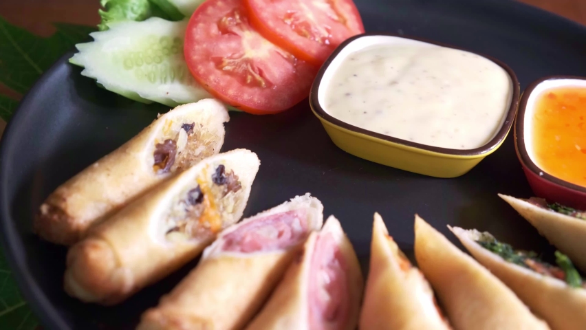 Delicious Spring Rolls with Dipping Sauce and Sliced Tomatoes and Cucumber on a Black Spinning Plate Close Up.