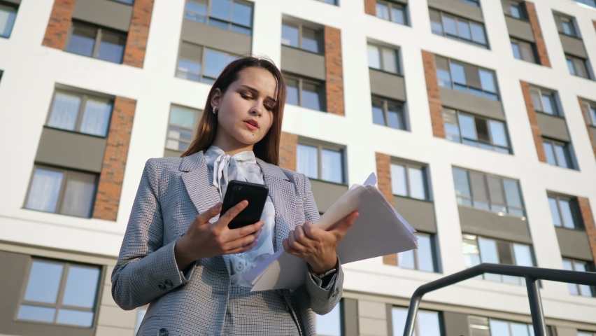 Young woman in a business suit looks at the phone and at the documents on the background of the building, slow motion. Royalty-Free Stock Footage #1078620167