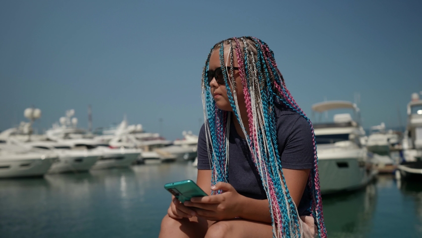 Fashionable teen girl with afro pigtails sitting on the shore of the seaport with a phone and sunglasses