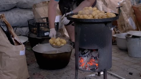 Street Chef Prepares Potatoes on a Wood Stove with Glowing Coals. Man shifts washed potatoes in a box made of cast iron cauldron. Cooking of grilled vegetables with steam and smoke. Zoom. Close up.