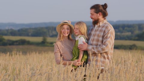 Happy caucasian family stands in wheat field in nature outdoors mother holds little girl daughter father takes off hat from wife puts on baby laughing smiling talking having fun on summer day vacation