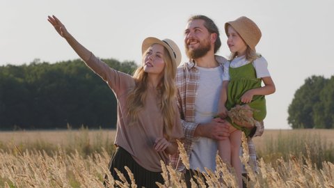 Happy caucasian family loving parents with little daughter child standing in wheat field in nature summer day, father holding small girl showing with finger direction talking, vacations outdoors
