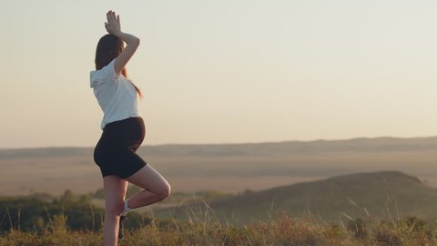 White pregnant woman in sports clothes is doing yoga outdoor, back view. Concept of mother and child health and meditation practices