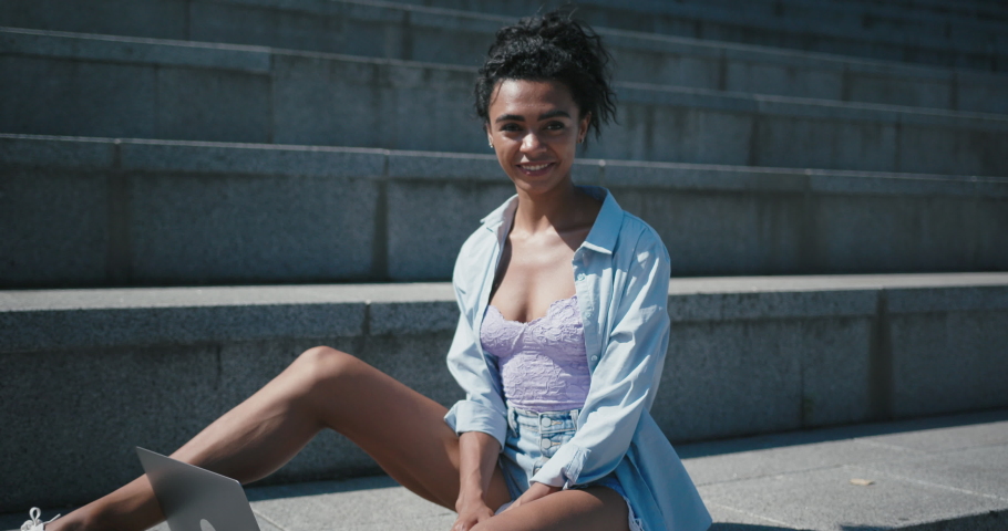 Attractive smiling African-American woman with bare legs sits by laptop on empty stone stairs in city on sunny summer day slow motion