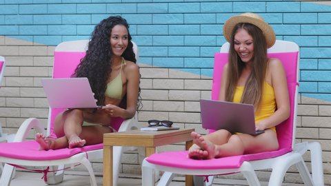 Two happy young multiracial Caucasian Hispanic women freelancers sitting on sun lounger relaxing at poolside and using laptops by pool while on summer vacation. Remote work anywhere in world concept