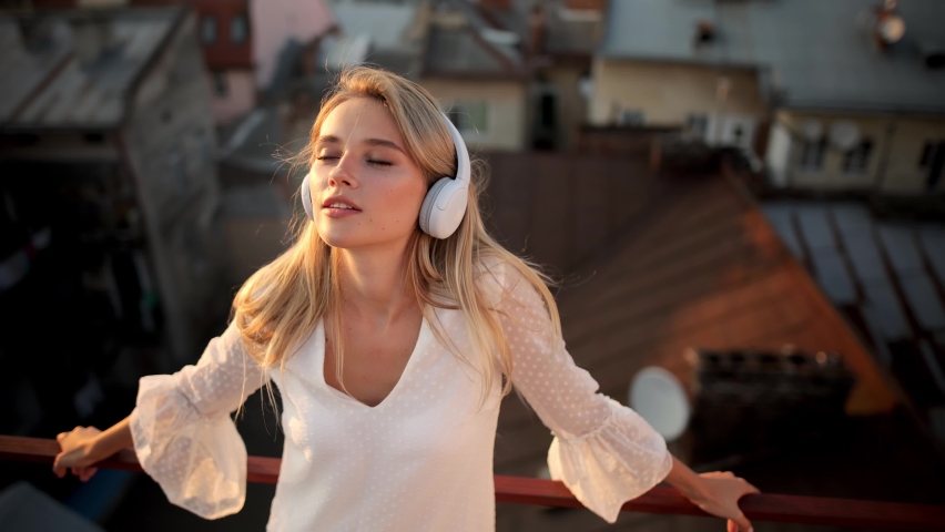 Peaceful blonde woman in modern wireless headphones relax on rooftop listening to music. Happy young girl in earphones rest on Roof Terrace at Sunset. Enjoy good quality sound. Stress free concept
