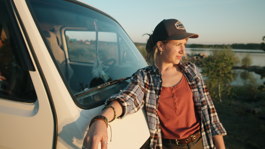 Zoom in shot of young beautiful woman leaning on van, looking at camera and smiling while standing at lakeside during golden hour on summer evening