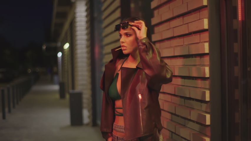 Party girl laughing urban background of modern night city. Fashion woman holding match in her teeth. Beautiful model wearing brown jacket, green bra, sunglasses. 4K