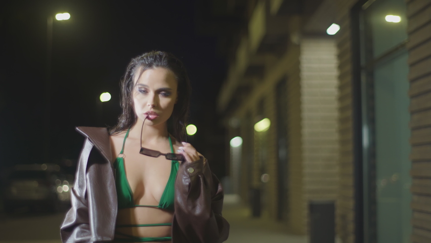 Sad woman holding sunglasses in night street. Stylish young beautiful model in brown jacket and green bra posing over night city background. 4K