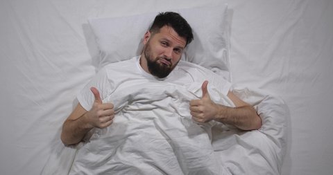 Top view of handsome bearded guy lying in bed with white linens in light room. High angle shooting. Man is looking down under the blanket and showing, thumbs up. Male health concept