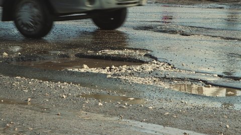 Cars drive in close-up on a bad, bumpy road with holes. Large potholes after rain, damaged road infrastructure. Asphalt in violation of technology. Taxpayer money is wasted and corruption is rampant.