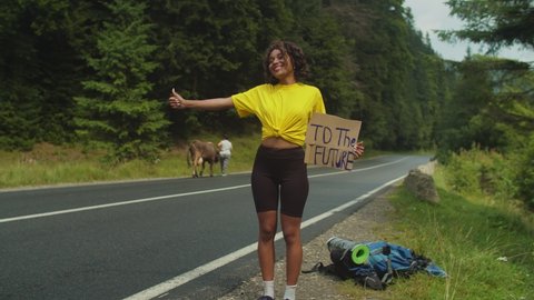 Portrait of charming smiling african american woman hiker with thumbs up and cardboard, with text to the future written on it, hitchhiking at mountain road while backpacking on summer vacations.