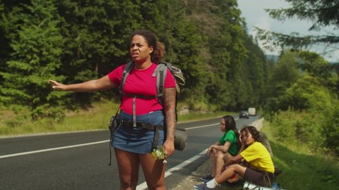 Tired pretty african american female traveler with backpack standing at roadside, hitchhiking with outstretched hand during mountain hiking with exhausted multiracial girlfriends resting in background