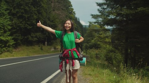 Lovely cheerful asian female tourist with backpack standing on roadside ,hitchhiking and signing thumb up for passing car, looking with radiant smile while enjoying mountain hiking in summertime.
