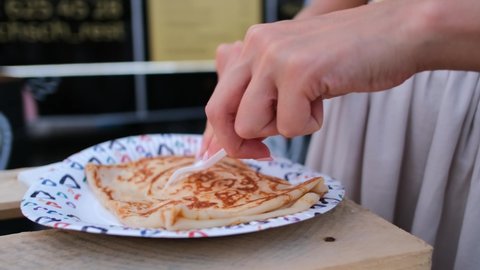 Woman hands cut big pancake with plastic knife and fork on coloured paper plate on wooden table in outdoor street cafe closeup