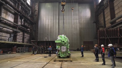 KAZAN, TATARSTAN RUSSIA - MAY 27 2021: Workers team controls carrying contemporary ship motor by crane in spacious dry repairing dock at industrial complex on May 27 in Kazan