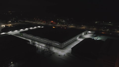 Motion around large illuminated storehouse with flat roof on snowy territory of production plant at night bird eye view