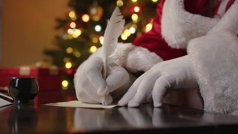 Santa Claus writes letter with quill pen on desk close-up with Happy New Year or Christmas greetings.