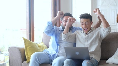 Happy excited grown son and mature 50s father using laptop, celebrating success. Two family generations of sport fan watching online game or match on computer, making yes win gesture, having fun
