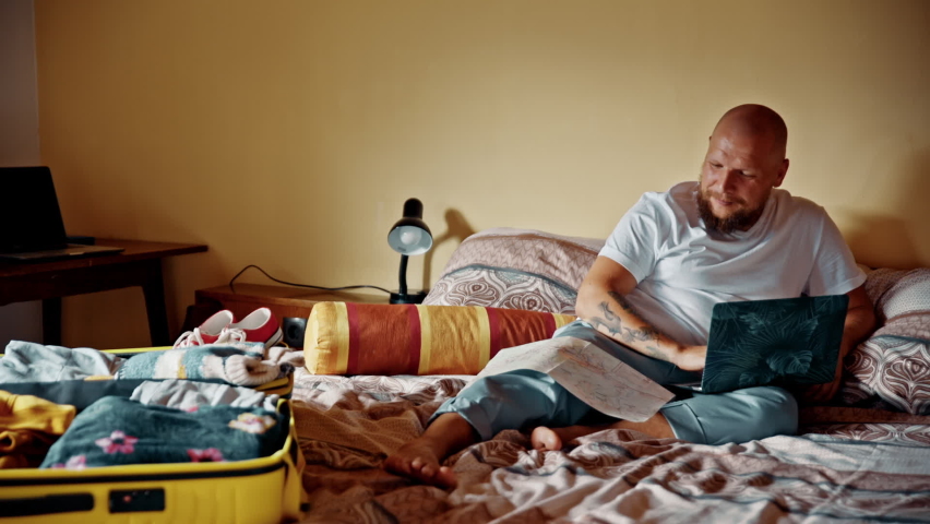 Man laying in bed with laptop preparing for vacation. Male smiling looking for hotel to stay and organizing route for upcoming trip. Concept of removal of restrictions, planning, travel.