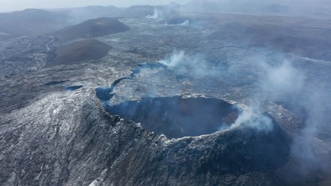 Otherworldly aerial drone view of smoky volcanic lake lava landscape, drone flying backwards reveals Fagradalsfjall crater cone, Iceland, sunny day