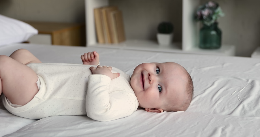 Close up of sweet beautiful newborn baby in bodysuit and diaper lying on bedsheets, plays touches toes laughs smile looks at camera. Carefree healthy babyhood, healthcare and paediatrics care concept Royalty-Free Stock Footage #1078681556