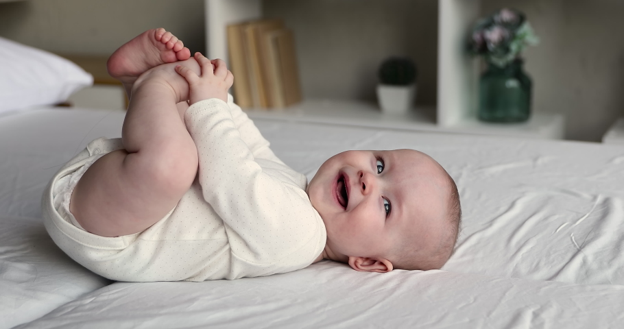 Close up of sweet beautiful newborn baby in bodysuit and diaper lying on bedsheets, plays touches toes laughs smile looks at camera. Carefree healthy babyhood, healthcare and paediatrics care concept Royalty-Free Stock Footage #1078681556